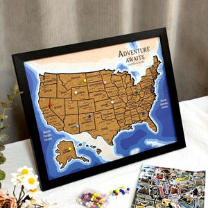 Pode incluir: Um mapa de cortiça emoldurado dos Estados Unidos com o texto Adventure Awaits United States. O mapa apresenta fronteiras estaduais, áreas oceânicas azuis e percevejos coloridos. Um pequeno recipiente transparente com percevejos e autocolantes.