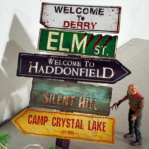 Könnte beinhalten: Ein vertikaler Wegweiser mit fünf rechteckigen und pfeilförmigen Schildern mit der Aufschrift Welcome to Derry, Elm St, Welcome to Haddonfield, Welcome to Silent Hill und Camp Crystal Lake Est 1935, begleitet von einer kleinen Freddy-Krueger-Figur.