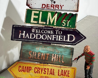 Peut inclure: Un poteau de signalisation vertical avec cinq panneaux rectangulaires et en forme de flèche portant les inscriptions Welcome to Derry, Elm St, Welcome to Haddonfield, Welcome to Silent Hill et Camp Crystal Lake Est 1935, avec une figurine.