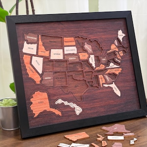 May include: A framed wooden puzzle of the United States, featuring individual state pieces in varying shades of brown and orange. The frame is black, and the background is a darker wood. Some state pieces are removed, and a few are scattered below.