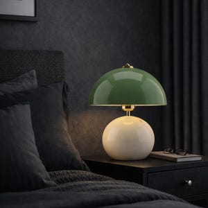 Könnte beinhalten: Eine grüne und cremefarbene Tischlampe mit rundem Schirm und Sockel. Die Lampe steht auf einem schwarzen Nachttisch neben einem Bett mit dunkelgrauer Bettwäsche. Eine Brille und ein Buch liegen ebenfalls auf dem Nachttisch.
