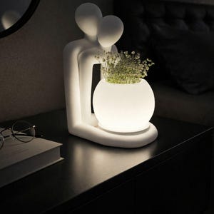Könnte beinhalten: Eine weiße Keramiklampe in Form von zwei sich umarmenden Figuren, die einen leuchtenden, kugelförmigen Pflanzentopf mit Grünzeug halten. Die Lampe steht auf einem dunklen Nachttisch und erzeugt ein warmes Umgebungslicht. Ein Buch und eine Brille liegen ebenfalls dort.
