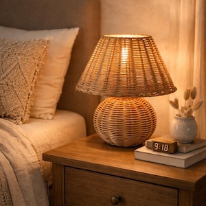 Natural rattan bedside table lamp featuring a woven wicker base and matching rattan lampshade that softly diffuses warm light. Placed on a wooden nightstand beside a bed, this lamp creates a cozy and calming bedroom atmosphere. The handcrafted wicker design adds texture and natural charm to interiors. Perfect for boho, farmhouse, coastal, or Scandinavian decor styles and ideal as a bedside or accent lighting piece.