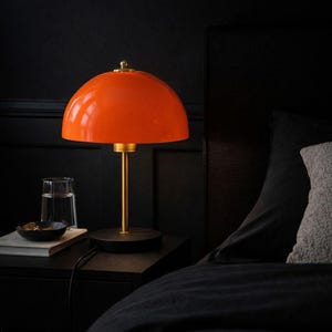Könnte beinhalten: Eine orangefarbene, kuppelförmige Tischlampe mit goldenem Stiel und Sockel steht auf einem schwarzen Nachttisch. Ein Glas Wasser und eine kleine Schale befinden sich ebenfalls auf dem Nachttisch. Die Lampe steht neben einem Bett mit schwarzer Bettwäsche.