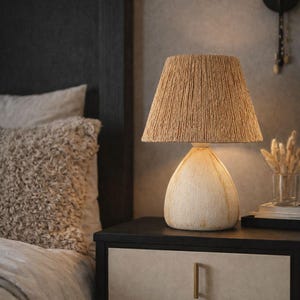 Könnte beinhalten: Eine beige Tischlampe mit strukturiertem, gewebtem Lampenschirm und cremefarbenem Sockel. Die Lampe steht auf einem schwarzen Nachttisch mit zwei Schubladen. Die Schlafzimmereinrichtung umfasst ein flauschiges Kissen und eine Wandleuchte.