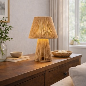 Könnte beinhalten: Eine Tischlampe mit gewebtem Schirm und Sockel. Die Lampe besteht aus einem hellbraunen, strukturierten Material und steht auf einer dunklen Holzoberfläche. Die Lampe ist beleuchtet und wirft einen warmen Schein. Eine kleine Schale und ein geflochtener Korb befinden sich ebenfalls auf dem Tisch.