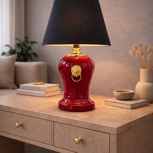 Luxury decorative table lamp with a red ceramic base and gold lion head ring accent paired with a classic black fabric lampshade. Displayed on a stylish console table, this lamp brings a bold and elegant statement to the interior. The warm lighting and rich colors create a sophisticated atmosphere. Ideal for living rooms, bedrooms, offices, or interiors with classic, modern luxury, or dark elegance decor styles.