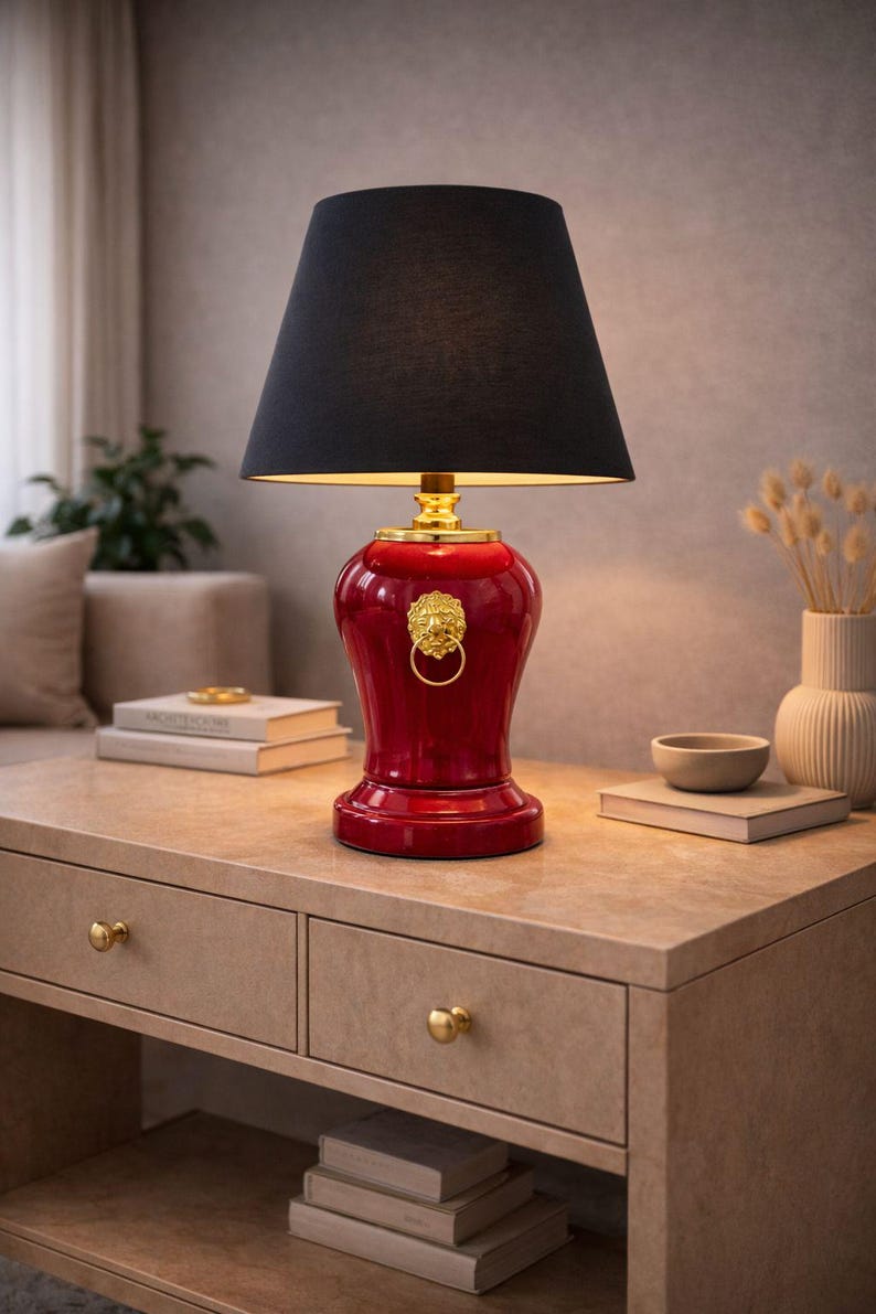 Luxury decorative table lamp with a red ceramic base and gold lion head ring accent paired with a classic black fabric lampshade. Displayed on a stylish console table, this lamp brings a bold and elegant statement to the interior. The warm lighting and rich colors create a sophisticated atmosphere. Ideal for living rooms, bedrooms, offices, or interiors with classic, modern luxury, or dark elegance decor styles.