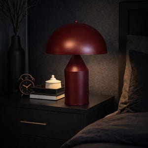 May include: A burgundy-colored table lamp with a rounded shade and cylindrical base sits on a black nightstand. A black vase with branches, a small alarm clock, and a decorative box are also on the nightstand. The scene suggests a bedroom setting.