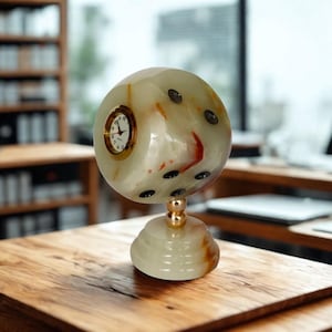 Könnte beinhalten: Eine einzigartige, kugelförmige Uhr aus cremefarbenem Onyx, die einem Würfel ähnelt. Das Zifferblatt ist goldfarben mit schwarzen Ziffern. Die Onyx-Kugel hat schwarze Punktmarkierungen und steht auf einem goldfarbenen Sockel. Die Uhr steht auf einer Holzoberfläche.