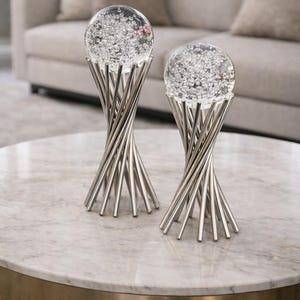 May include: Two decorative glass globe sculptures with a swirling metal base. The clear globes are filled with small, sparkling elements. The metal bases are silver and have a twisted design. The sculptures are on a marble-topped table.
