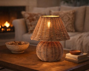 May include: A woven rattan table lamp with a conical shade and a rounded base. The lamp is lit, casting a warm glow. It sits on a wooden table, with a bowl of nuts and books nearby. The background includes a fireplace and a sofa.
