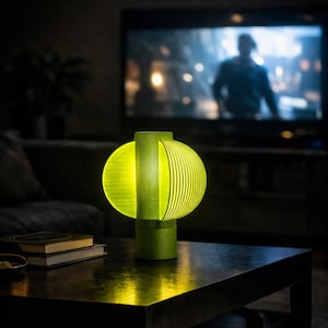 Könnte beinhalten: Eine moderne grüne Tischlampe mit einem kugelförmigen, geschlitzten Schirm, der ein warmes, gelbes Leuchten ausstrahlt. Die Lampe steht auf einem dunklen Holztisch, im Hintergrund ein Fernseher und Bücher. Das Design der Lampe ist modern und stilvoll.