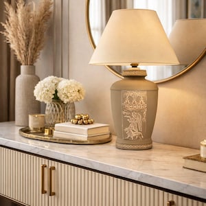 Könnte beinhalten: Eine beige Tischlampe mit cremefarbenem Lampenschirm. Der Lampenfuß hat ein dekoratives Design. Die Lampe steht auf einem Konsolentisch mit Marmorplatte und goldenen Akzenten. Dekorative Elemente sind eine Vase mit getrockneten Pflanzen, Blumen und Bücher.