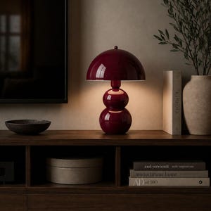 May include: A burgundy table lamp with a rounded shade and a stacked, spherical base. The lamp sits on a dark wood cabinet, next to books and a vase with greenery. A dark bowl and a box are also on the cabinet.
