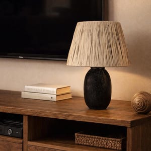 Könnte beinhalten: Dekorative Tischlampe mit strukturiertem beigefarbenem Lampenschirm und dunklem, ovalem Sockel. Die Lampe steht auf einer Holzoberfläche mit Büchern und einer dekorativen Kugel. Ein stilvolles Wohnaccessoire.