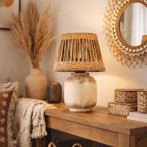 Könnte beinhalten: Dekorative Tischlampe mit gewebtem Rattanschatten und einem weißen, antik aussehenden Sockel. Die Lampe steht auf einem Holztisch, zusammen mit gewebten Körben und einem dekorativen Spiegel. Der Raum hat eine warme, neutrale Farbpalette.