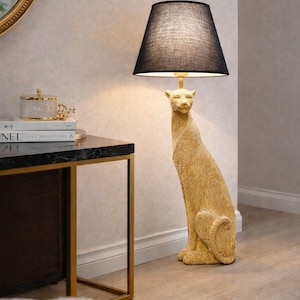 Könnte beinhalten: Eine goldfarbene Stehlampe in Form eines sitzenden Leoparden. Die Lampe hat einen schwarzen Lampenschirm und eine strukturierte Oberfläche. Die Lampe ist ein dekoratives Element für die Inneneinrichtung.