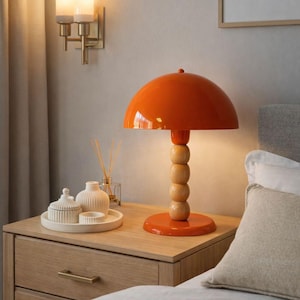 Könnte beinhalten: Orangefarbene Tischlampe in Pilzform mit Holzsockel und gestapelten Holzkugeln. Die Lampe steht auf einem Nachttisch aus Holz. Die Lampe ist etwa 40 cm hoch. Eine Wandleuchte mit zwei Lichtern ist sichtbar.