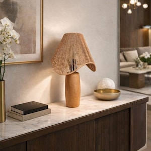 Könnte beinhalten: Eine Tischlampe aus Holz mit einem gewebten Lampenschirm. Die Lampe hat einen hellbraunen Holzsockel und einen beigefarbenen gewebten Schirm. Die Lampe steht auf einem Tisch mit Marmorplatte und einem dunkelbraunen Holzschrank. Links steht eine goldene Vase mit weißen Blumen.