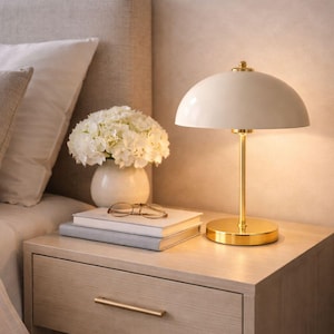 Könnte beinhalten: Eine Nachttischszene mit einer cremefarbenen Kuppellampe mit goldenem Sockel und Stiel, die auf einem Nachttisch aus hellem Holz steht. Eine Vase mit weißen Blumen, Bücher und eine Brille befinden sich ebenfalls auf dem Nachttisch.