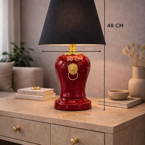 Decorative red ceramic table lamp with black fabric lampshade shown with size reference measuring 48 cm height and 30 cm width. The lamp features a gold lion head ring detail that enhances its luxurious appearance. This stylish lighting piece blends classic design with modern interiors. Ideal as a bedside lamp, console lamp, or accent lighting piece for living rooms, bedrooms, and elegant home decor spaces.
