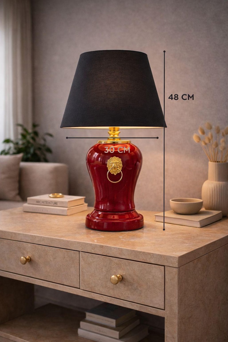 Decorative red ceramic table lamp with black fabric lampshade shown with size reference measuring 48 cm height and 30 cm width. The lamp features a gold lion head ring detail that enhances its luxurious appearance. This stylish lighting piece blends classic design with modern interiors. Ideal as a bedside lamp, console lamp, or accent lighting piece for living rooms, bedrooms, and elegant home decor spaces.