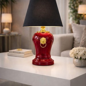 Elegant red ceramic table lamp with a glossy decorative base and black fabric lampshade displayed on a modern white console table. The lamp features a gold lion head ring handle detail that adds a luxurious and classic accent. The combination of deep red ceramic and black shade creates a sophisticated contrast while providing warm ambient lighting. Perfect for living rooms, entryway consoles, or luxury interior decor styles.