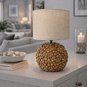 May include: A decorative table lamp with a beige woven shade and a round base covered in small, gold-colored accents. The lamp sits on a white surface, with a candle in a glass holder nearby.