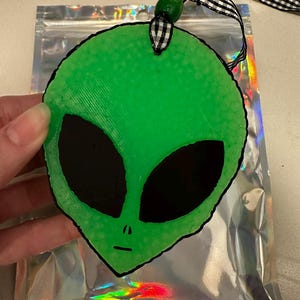 May include: A green alien head ornament with large black eyes and a small mouth. The ornament is attached to a black and white checkered ribbon and a green bead. The ornament is held in front of a silver holographic bag.