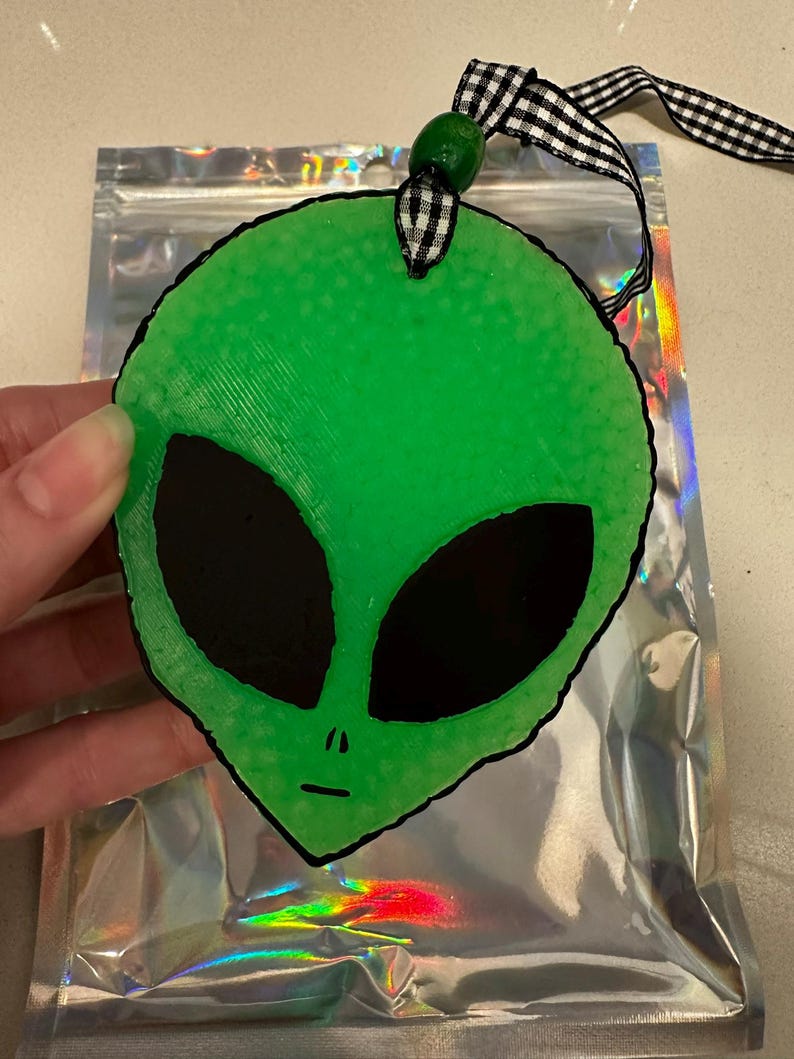 May include: A green alien head ornament with large black eyes and a small mouth. The ornament is attached to a black and white checkered ribbon and a green bead. The ornament is held in front of a silver holographic bag.