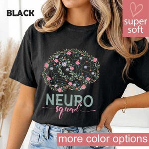 May include: Black t-shirt with a floral brain design and the words "NEURO squad". The design features pink and white flowers and green leaves. The shirt also has the words "more color options" in a pink banner.