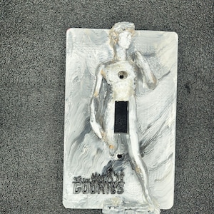 May include: A decorative light switch cover featuring a sculpted figure in a classical pose. The cover is painted in shades of gray and white with a marbled effect. The text "The Goonies" is printed at the bottom.