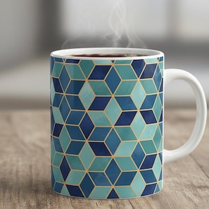 May include: White ceramic mug featuring a geometric design of blue and teal diamonds with gold outlines. The mug contains a dark liquid, with steam rising. The mug is on a wooden surface.