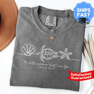 Könnte beinhalten: Ein graues Comfort Colors T-Shirt mit einer weißen Strichzeichnung einer Muschel, einer Schildkröte und einem Seestern. Der Text "Be still and know that I am God, Psalm 46:10" ist darunter gedruckt. Eine silberne Halskette mit Anhänger ist ebenfalls sichtbar.