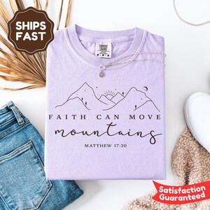 May include: A light purple t-shirt with the text "Faith Can Move Mountains" and a mountain graphic. The shirt is folded and has a silver necklace on top. The text "Matthew 17:20" is below the main text.