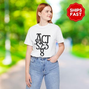 Op de afbeelding: Wit T-shirt met de tekst "ACT 2:3 8" in een zwarte, gestileerde letter. Het shirt wordt gecombineerd met een blauwe jeans. Een rood-witte "SHIPS FAST" sticker is rechtsboven zichtbaar.