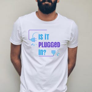 Puede incluir: Camiseta blanca con el texto "IS IT PLUGGED IN?" en azul y morado. El diseño incluye un gráfico estilizado de enchufe y toma de corriente. La camiseta es de manga corta y parece ser de algodón.