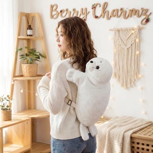 May include: A white plush seal backpack featuring a soft, furry texture, black button eyes, and a small nose. The backpack has adjustable straps and is worn on the back, designed in the shape of a full-body seal with flippers and a rounded head.