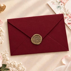 May include: A deep red envelope sealed with a gold wax stamp featuring the text Thank You. The envelope is surrounded by light pink roses, white baby's breath flowers, a gold wax seal stamp tool, and a wedding invitation card.