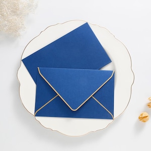 May include: Two deep blue envelopes with a smooth matte finish. One envelope is closed with a pointed flap featuring a thin gold metallic border along the edges, while the other is positioned behind it as a flat rectangular piece of blue stationery.