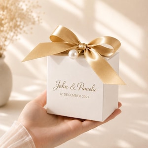 Op de afbeelding: Een vierkante witte geschenkdoos met een gouden satijnen strik bovenop, versierd met een enkele witte parel. Op de voorzijde van de doos staat de tekst John & Pamela en de datum 12 DECEMBER 2027 in een gouden script en schreefloos lettertype.