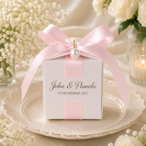 May include: A square white gift box featuring a light pink satin ribbon tied in a bow at the top with a single white pearl bead accent. The front of the box is printed with the names John & Pamela and the date 12 DECEMBER 2027 in black text.