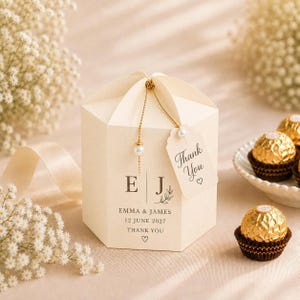 Può includere: Una scatolina esagonale color crema con le iniziali E e J, i nomi Emma & James e la data 12 giugno 2027. Una catenella dorata con una perla fissa un cartellino con la scritta Thank You e un piccolo cuore sulla parte superiore della scatola.