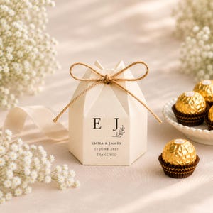 Può includere: Una scatolina esagonale color crema chiusa con un fiocco di spago marrone. Sul davanti sono stampate le lettere E e J, una piccola illustrazione di una foglia e il testo Emma & James, 12 giugno 2027, Thank You. Cioccolatini dorati sono vicini.