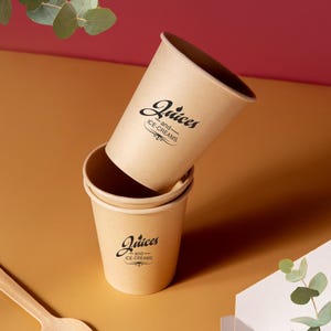 Op de afbeelding: Drie kraftpapieren bekers, twee gestapeld en één gekanteld, met de woorden "Juices and Ice-Creams" in zwart schrift. De bekers staan op een warmgekleurd oppervlak met een houten lepel in de linkerbenedenhoek.