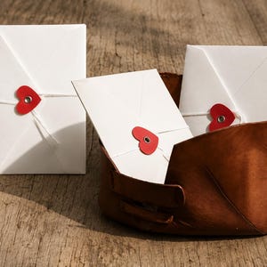 May include: Three white rectangular envelopes secured with white string and a red heart-shaped closure featuring a central metal grommet. Two envelopes are tucked inside a brown leather bowl, while one stands upright to the left of the bowl.