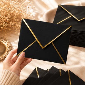 May include: A rectangular black envelope featuring a pointed flap with a metallic gold border. The envelope is held in a hand, with a stack of identical black envelopes with gold trim visible in the background and foreground of the composition.