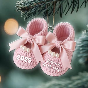 Op de afbeelding: Een paar lichtroze gehaakte babyslofjes als kerstornament. Elke slof is voorzien van een satijnen strik, drie witte imitatieparels en witte tekst. Op de ene slof staat 2025, op de andere staat Laura's first step.