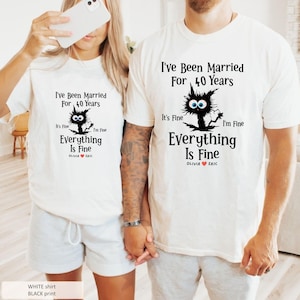 May include: Two white t-shirts featuring a black graphic of a messy-haired cat with large blue eyes. The text reads: I've Been Married For 40 Years, It's Fine I'm Fine, Everything Is Fine, and the names Olivia and Eric with a red heart symbol.
